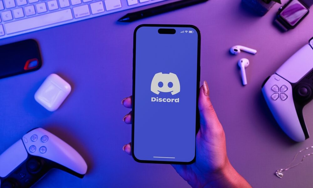 Fille tenant un smartphone iPhone 14 Pro avec application Discord avec contrôleurs de jeux vidéo PS5. Vue de dessus. Environnement de bureau. Basse lumière bleue. @Shutterstock
